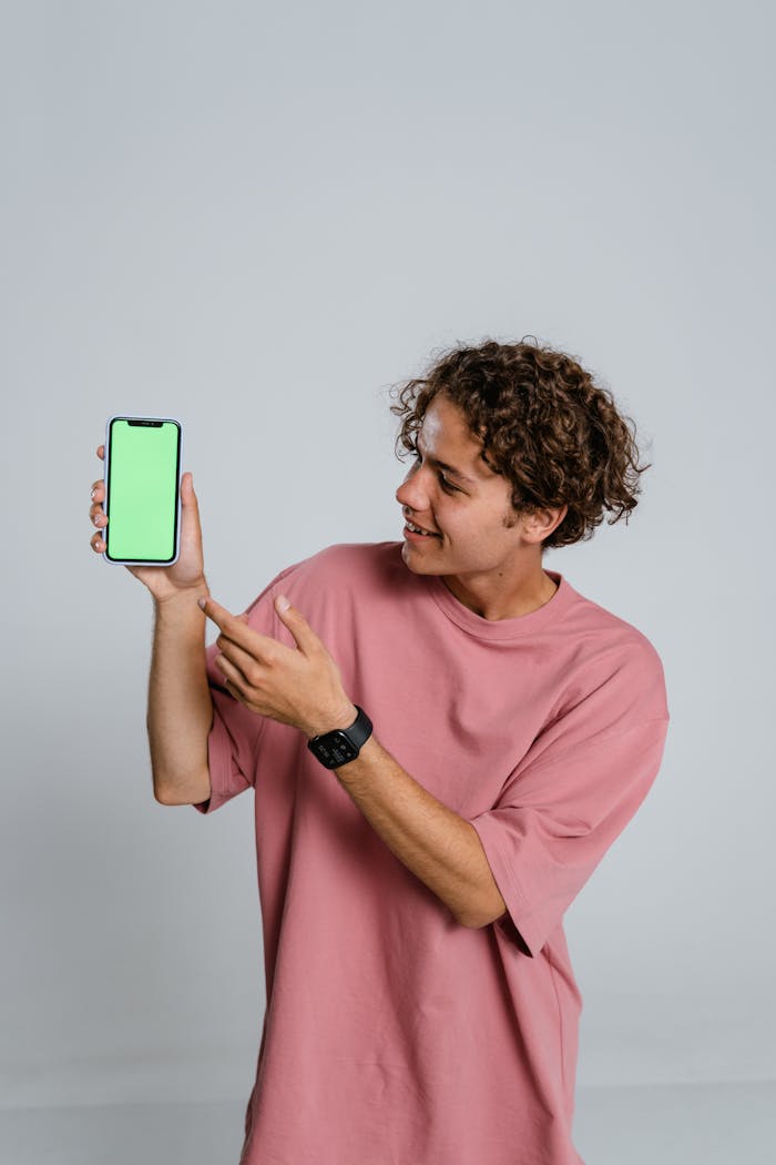 Smiling young man in pink shirt holding a smartphone with a green screen against a neutral background.